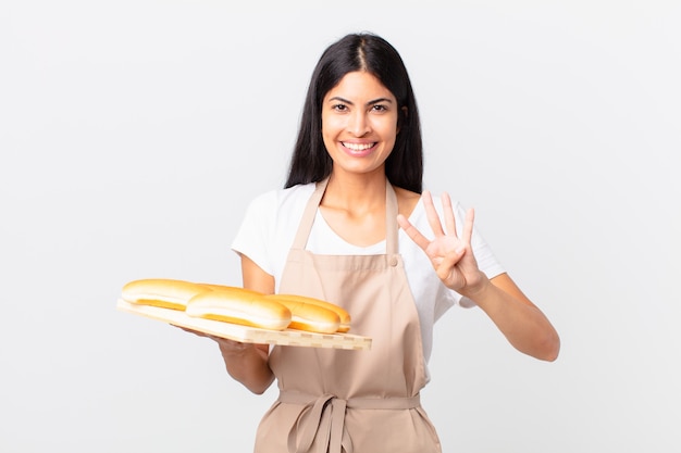 Mujer bonita chef hispana sonriendo y mirando amigable, mostrando el número cuatro y sosteniendo una bandeja con bollos de pan