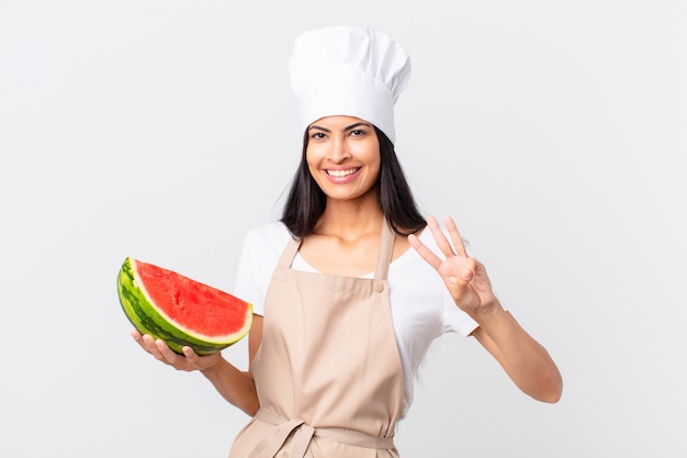 Mujer bonita chef hispana sonriendo y mirando amigable, mostrando el número tres y sosteniendo una sandía