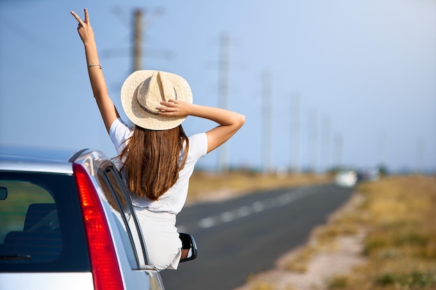 Mujer bonita delgada con sombrero de paja disfrutando de un viaje por carretera en un día de verano Mujer joven emocionada levantando la mano con el signo de la victoria de la ventana del auto Chica sentada en la puerta del auto y asomándose por la ventana