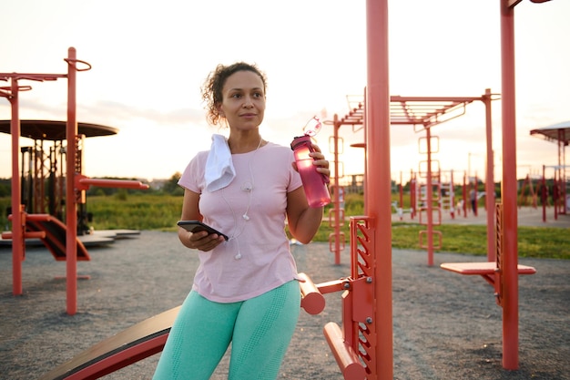 Mujer bonita descansa en el campo de deportes después de hacer ejercicio en máquinas de gimnasio sostiene una botella con agua y un teléfono inteligente y mira hacia otro lado contra el fondo del atardecer