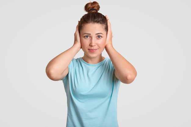 Una mujer bonita y desesperada con un nudo en el pelo mantiene las manos en las orejas, ignora el sonido fuerte, se viste con una camiseta informal, posa contra la pared blanca. Concepto de personas y sentimientos