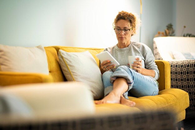 Una mujer bonita disfruta relajándose en casa leyendo un libro sobre el lector de tabletas y bebiendo un té o un café