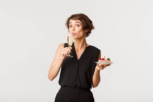 Mujer bonita divertida bebiendo champán y sosteniendo un trozo de pastel de cumpleaños, de pie sobre blanco.