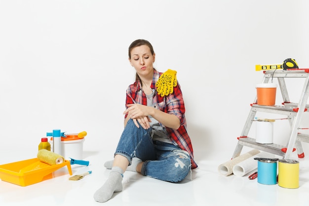 Mujer bonita divertida en ropa casual sentada en el piso con guantes amarillos, instrumentos para apartamento de renovación aislado sobre fondo blanco. Accesorios de papel pintado para herramientas de pintura. Reparar el concepto de hogar