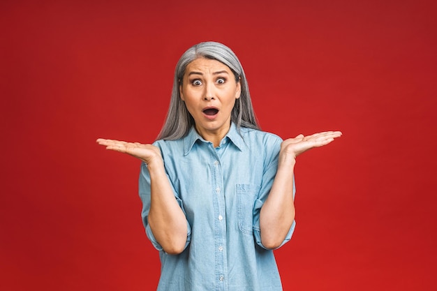 Mujer bonita de edad avanzada que se siente sorprendida y emocionada riéndose asombrada y feliz debido a una sorpresa inesperada aislada sobre un fondo rojo