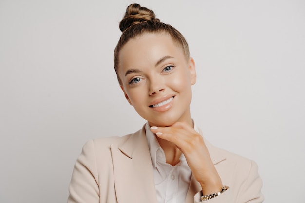 Mujer bonita ejecutiva en traje beige y cabello en moño en la conferencia de negocios escuchando buenas noticias, siendo feliz y relajado mientras mira a la cámara con la mano en la barbilla aislada sobre fondo gris