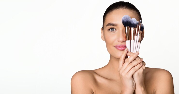 Mujer bonita escondiendo la mitad de la cara detrás de pinceles de maquillaje posando sobre un fondo gris