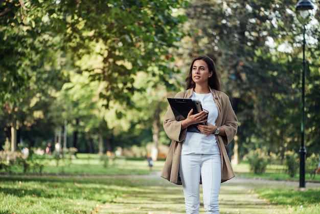 Mujer bonita está de pie con la carpeta portátil y la computadora portátil en el fondo del parque de verano