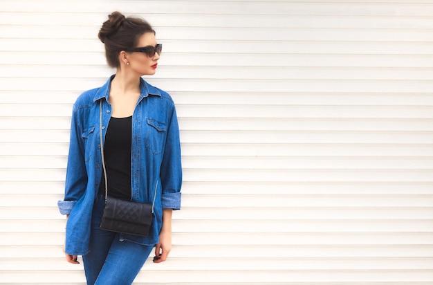 Foto mujer bonita con estilo en gafas de sol con una pequeña bolsa en conjunto de mezclilla de pie cerca de la pared blanca