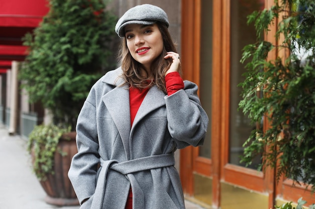Mujer bonita con estilo joven con vestido rojo abrigo gris y sombrero posando en las calles de la ciudad