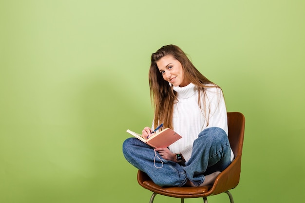 Mujer bonita europea en suéter blanco casual aislado, lindo feliz sentarse en una silla con bloc de notas y bolígrafo