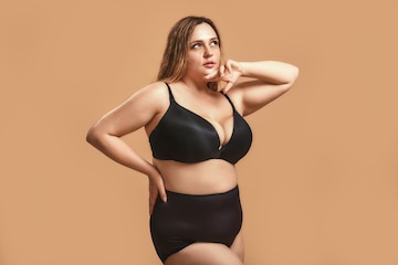 Mujer bonita gordita en lencería negra sexy a otro lado está de pie contra el marrón en el estudio. con sobrepeso | Foto Premium