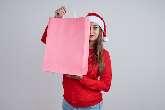 Una mujer bonita con un gorro de Papá Noel, con un suéter rojo, escondida detrás de bolsas de la compra. El concepto de ventas navideñas, compras, descuentos.