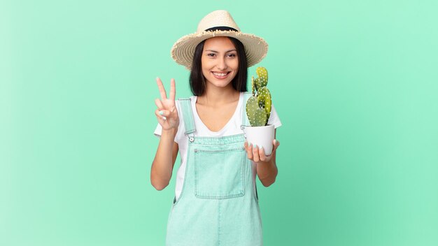 Mujer bonita granjera sonriendo y mirando amigable, mostrando el número dos y sosteniendo un cactus