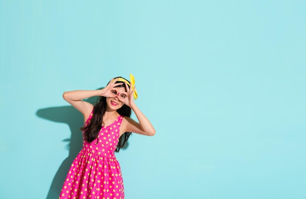 una mujer bonita hace un gesto bien para cubrirse los ojos y muestra la ropa encantadora con un vestido rosa y una banda de pelo amarilla en el fondo azul de la pared con un área en blanco sobre el espacio de copia.