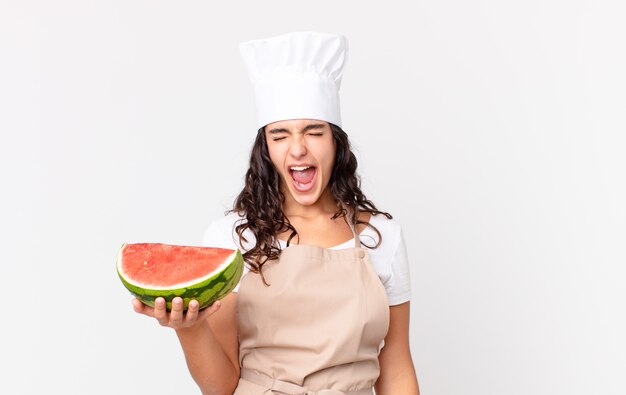 Mujer bonita hispana chef gritando agresivamente, mirando muy enojado y sosteniendo una sandía