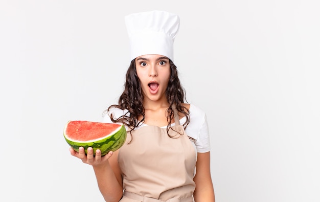 Mujer bonita hispana chef mirando muy conmocionado o sorprendido y sosteniendo una sandía