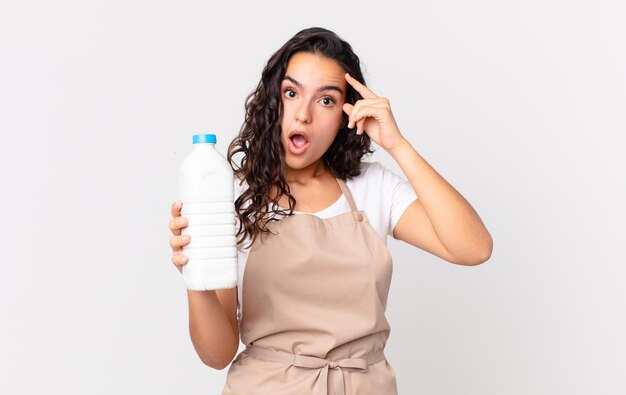 Mujer bonita hispana chef mirando sorprendida, dándose cuenta de un nuevo pensamiento, idea o concepto y sosteniendo una botella de leche