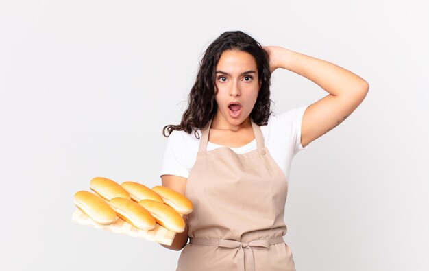 Mujer bonita hispana chef sintiéndose estresada, ansiosa o asustada, con las manos en la cabeza y sosteniendo un pan troy