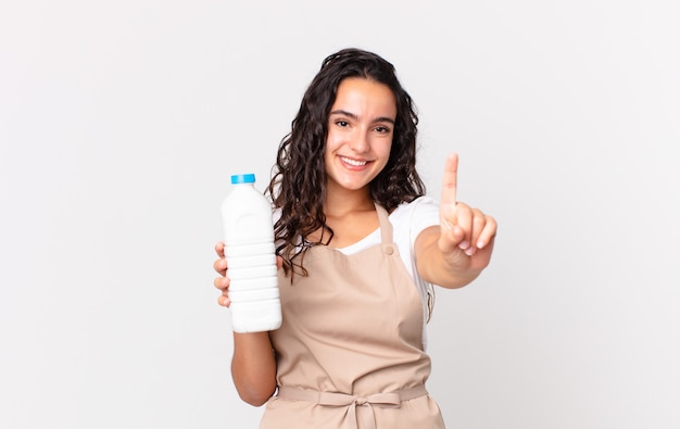 Mujer bonita hispana chef sonriendo y mirando amigable, mostrando el número uno y sosteniendo una botella de leche