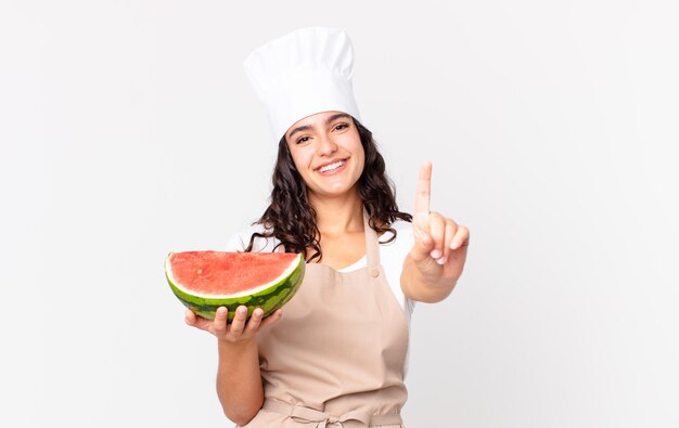 Mujer bonita hispana chef sonriendo y mirando amigable, mostrando el número uno y sosteniendo una sandía