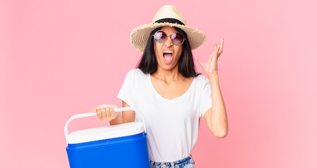 Mujer bonita hispana gritando con las manos en el aire con un refrigerador portátil de picnic