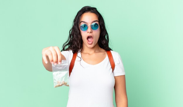 Mujer bonita hispana mirando muy conmocionado o sorprendido y sosteniendo una bolsa de marihuana