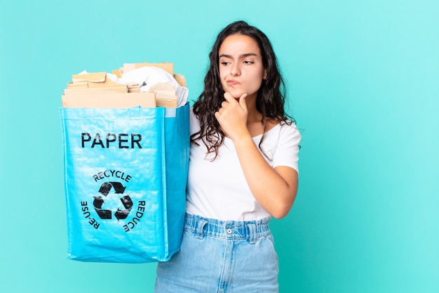 Mujer bonita hispana pensando, sintiéndose dudoso y confundido y sosteniendo una bolsa de papel reciclado