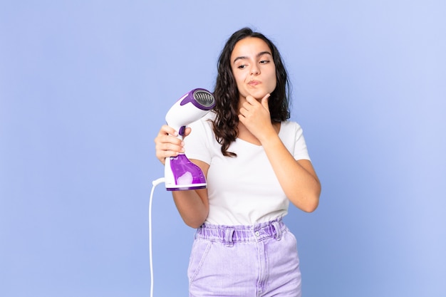 Mujer bonita hispana pensando, sintiéndose dudoso y confundido y sosteniendo una plancha de vapor