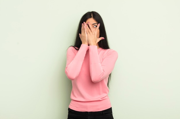 Mujer bonita hispana que cubre la cara con las manos mirando entre los dedos con expresión sorprendida y mirando hacia un lado