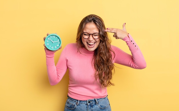 Mujer bonita hispana que parece infeliz y estresada, gesto de suicidio haciendo signo de pistola. concepto de reloj despertador