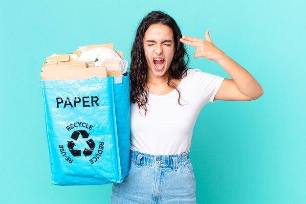 Mujer bonita hispana que parece infeliz y estresada, gesto de suicidio haciendo un signo de pistola y sosteniendo una bolsa de papel reciclado