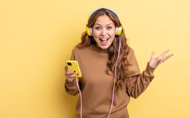 Mujer bonita hispana que se siente feliz y asombrada por algo increíble concepto de auriculares y teléfono inteligente