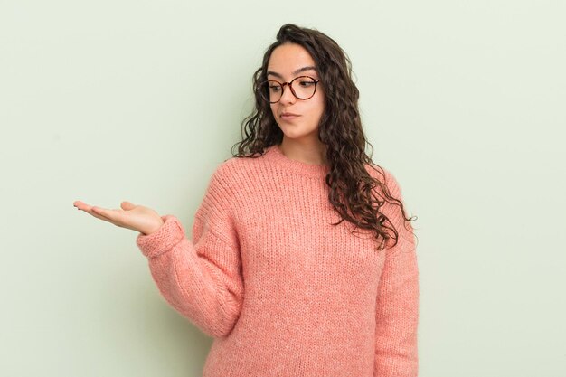 Mujer bonita hispana que se siente feliz y sonriendo casualmente mirando un objeto o concepto sostenido en la mano al costado