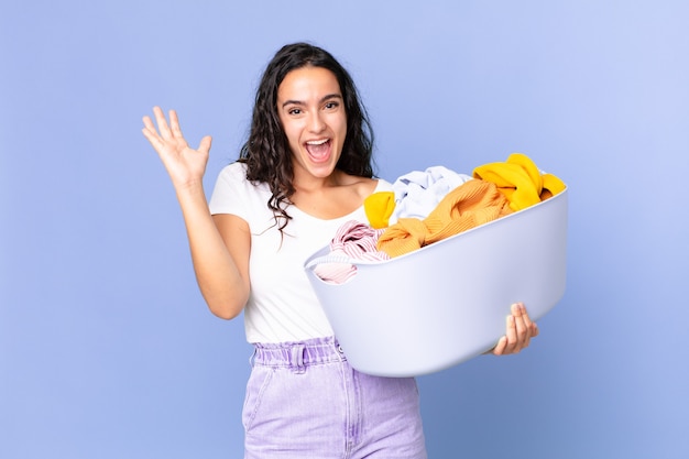 Mujer bonita hispana sintiéndose feliz y asombrada por algo increíble y sosteniendo una canasta de lavar ropa