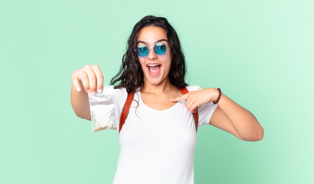 Mujer bonita hispana sintiéndose feliz y señalando a sí misma con un emocionado y sosteniendo una bolsa de marihuana