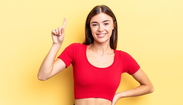 Mujer bonita hispana sonriendo alegre y felizmente, apuntando hacia arriba con una mano para copiar espacio