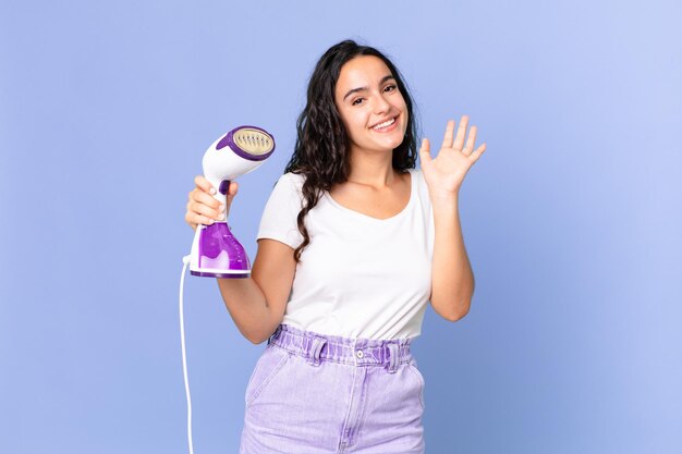 Mujer bonita hispana sonriendo alegremente, saludando con la mano, dándote la bienvenida y saludándote y sosteniendo un vapor de hierro