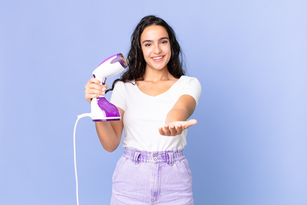 Mujer bonita hispana sonriendo felizmente con amable y ofreciendo y mostrando un concepto y sosteniendo una plancha de vapor