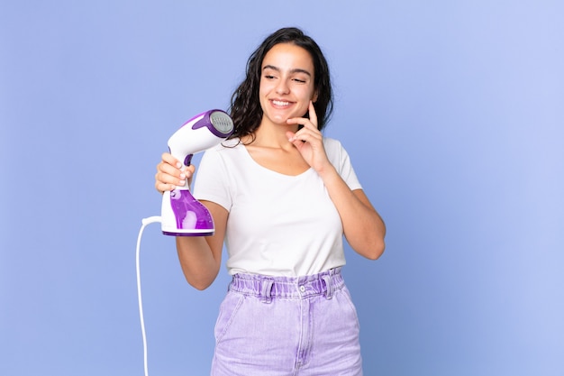 Mujer bonita hispana sonriendo felizmente y soñando despierto o dudando y sosteniendo una plancha de vapor