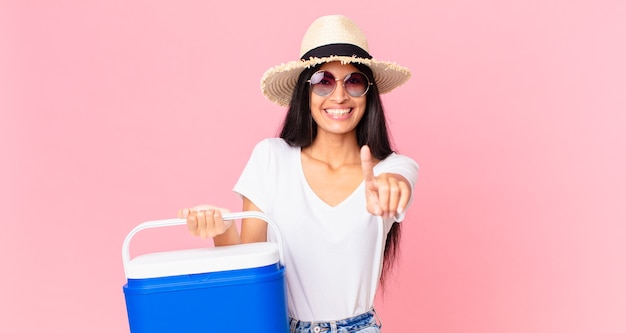 Mujer bonita hispana sonriendo con orgullo y confianza haciendo el número uno con un refrigerador portátil de picnic