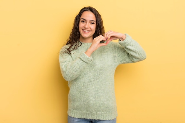 Mujer bonita hispana sonriendo y sintiéndose feliz linda romántica y enamorada haciendo forma de corazón con ambas manos