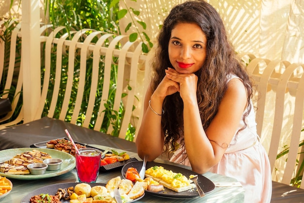 Mujer bonita india con vestido rosa y peinado rizado sentada en un café tropical de verano