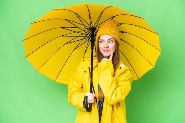 Mujer bonita joven con abrigo impermeable y paraguas sobre un fondo aislado de clave de croma pensando en una idea mientras mira hacia arriba