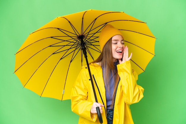 Mujer bonita joven con abrigo impermeable y paraguas sobre fondo de clave de croma aislado gritando con la boca abierta a un lado