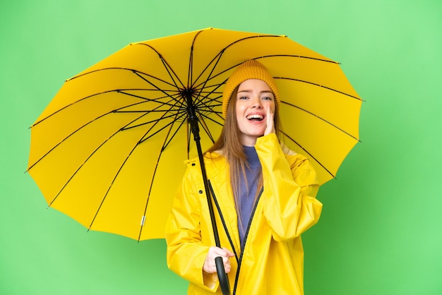 Mujer bonita joven con abrigo impermeable y paraguas sobre fondo de clave de croma aislado gritando con la boca abierta