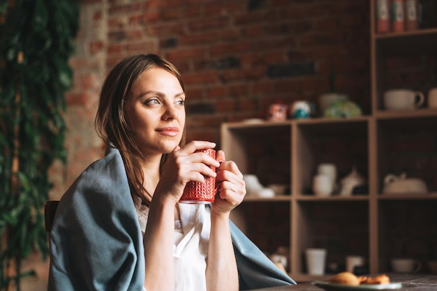 Mujer bonita joven en acogedora bufanda gris con taza de té en las manos mira por la ventana y descansa en su estudio