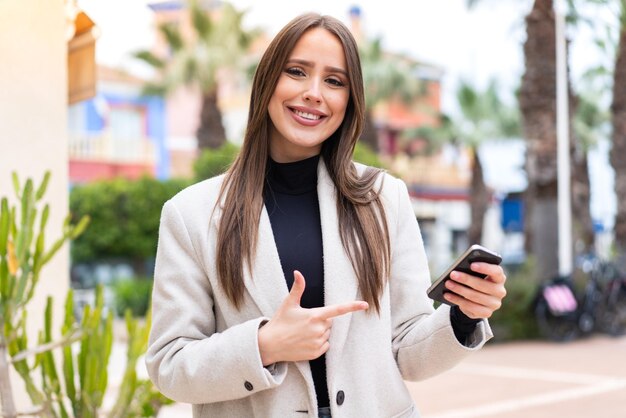 Mujer bonita joven al aire libre mediante teléfono móvil y apuntando