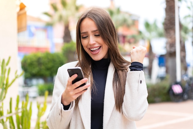 Mujer bonita joven al aire libre con teléfono móvil y haciendo el gesto de la victoria