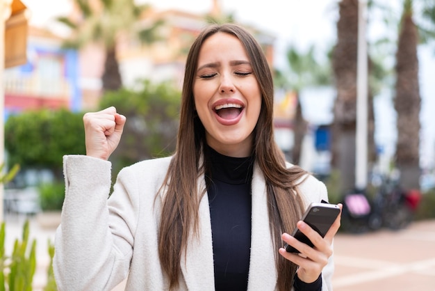 Mujer bonita joven al aire libre con teléfono móvil y haciendo el gesto de la victoria